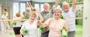Dans une salle de sport, deux femmes âgées aux cheveux blonds et deux hommes âgés aux cheveux blancs tiennent des tapis d'exercice verts et envoient la main en souriant