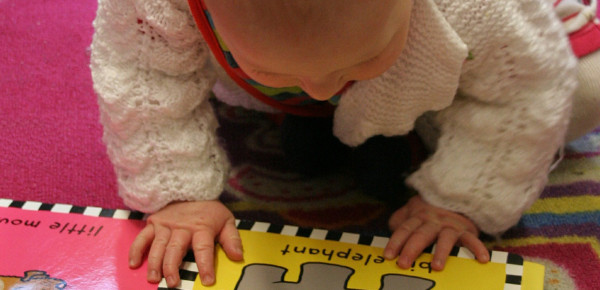Sur un tapis rose, un bébé avec un chandail de laine blanc lit un livre avec des images d'animaux