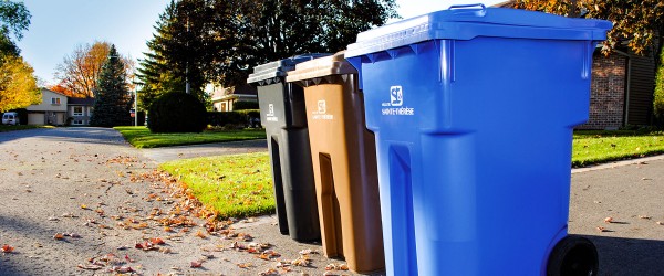 En bordure de rue au soleil, un bac de recyclage bleu, un bac de compost brun et bac noir à ordures sont alignés
