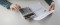 Sur une table blanche, une personne tient une feuille dans une main et tape sur une calculatrice avec son autre main