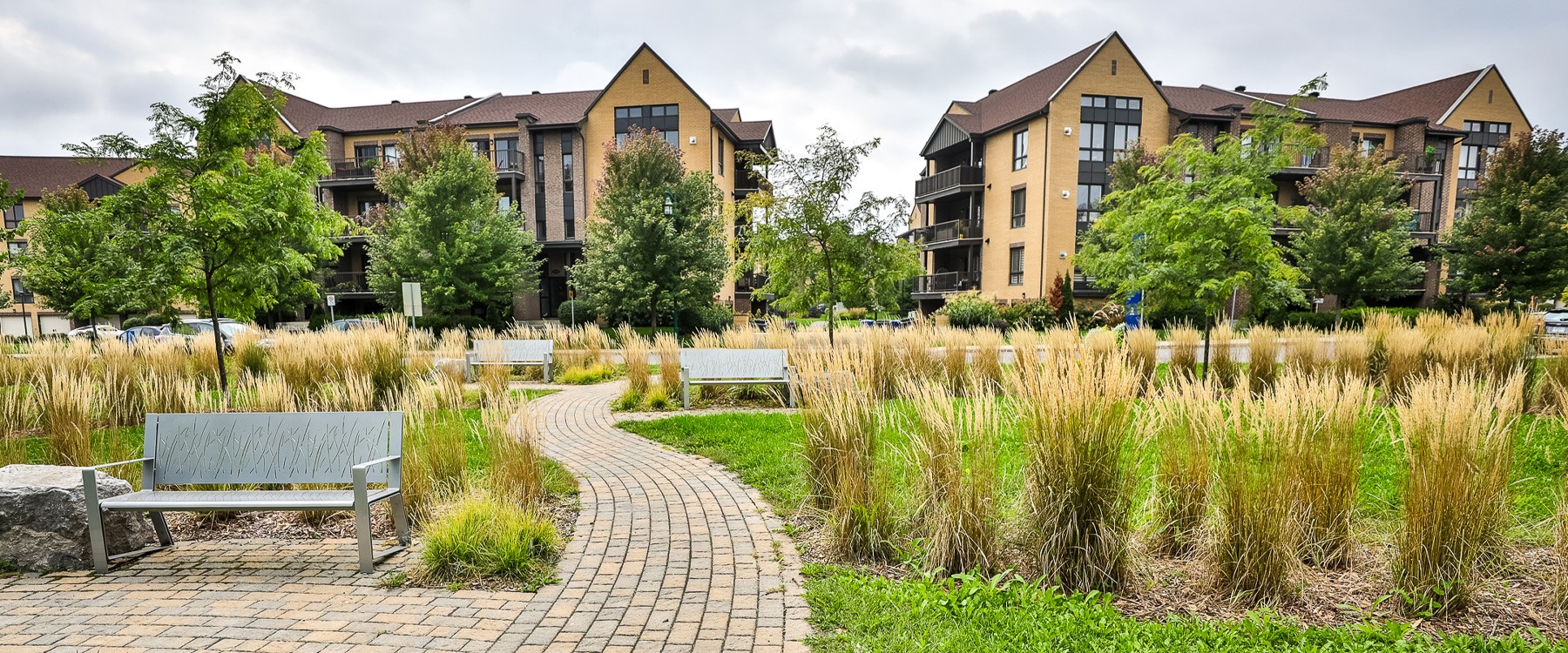 Sentier en pavé uni qui serpente à travers un parc. Trois bancs de parcs en métal sont installés en bordure et des immeubles avec des logements se trouvent en arrière-plan