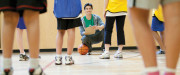 Dans un gymnase, des jeunes en habits de sport sont debout devant un enseignant accroupi avec un calepin de notes et un ballon de basketball