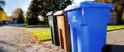En bordure de rue au soleil, un bac de recyclage bleu, un bac de compost brun et bac noir à ordures sont alignés côte à côte