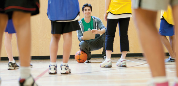 Dans un gymnase, des jeunes en habits de sport sont debout devant un enseignant accroupi avec un calepin de notes et un ballon de basketball