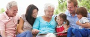 Assis à l'extérieur côte à côte, une famille composée de deux parents, deux jeunes enfants et deux grands-parents regardent un livre en souriant