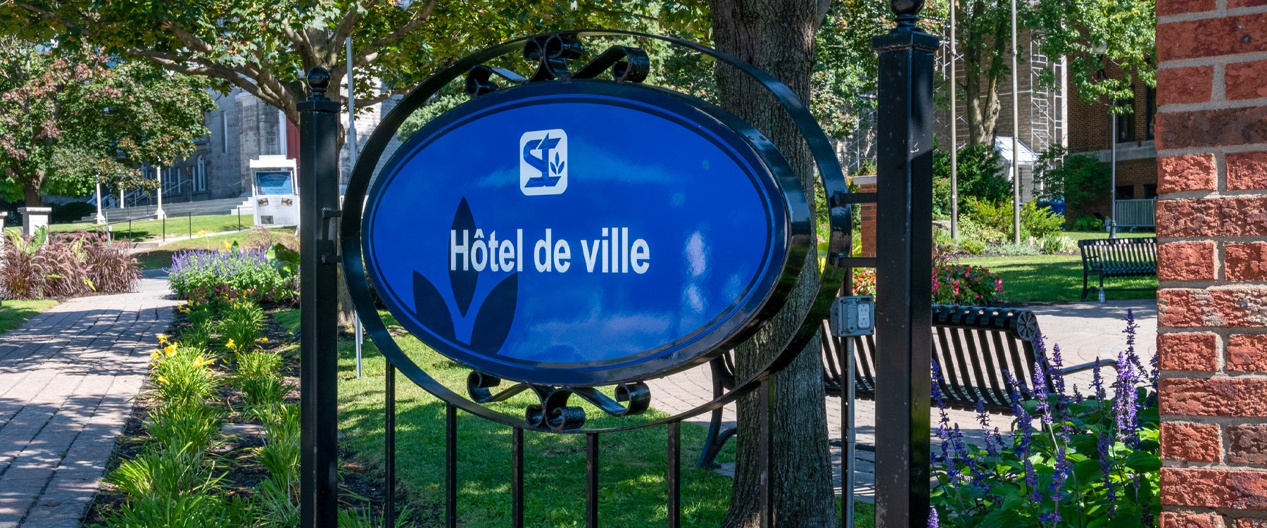 À l'extérieur sous les arbres, clôture en fer noir avec écriteau bleu avec le logo de la Ville et l'inscription hôtel de ville