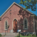 Façade avant d'une église de briques rouges avec deux fenêtres en pointe, une porte haute en bois et des escaliers en béton menant à l'entrée
