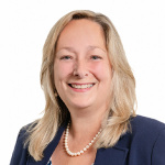 Femme blanche aux cheveux blonds porte un tailleur bleu marin et un collier de perles blanches