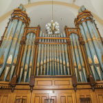 Dans une église, grand orgue fait de bois orangé dont les tuyaux sont peinturés en turquoise avec des ornements en or.