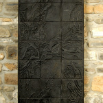 Œuvre rectangulaire composée de 27 carrés de céramique texturés noirs collés ensemble et accrochés sur un mur de pierre.