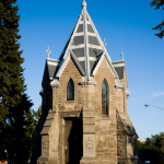 Petite chapelle circulaire faite de pierres grises avec une noire et des fenêtres en vitrail. Le toit gris et blanc monte en pointe.