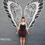 Sur un mur de briques grises, une femme blanche aux cheveux roux portant une robe courte noire se tient debout devant une murale de peinture en forme d'ailes noires et blanches