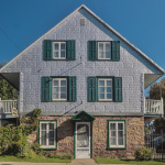 Façade d'une maison faite en briques blanches sur le haut et en pierre au rez-de-chaussée. La porte d'entrée est blanche et des volets verts accompagnent les six fenêtres.
