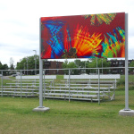 Installés dans un parc devant des estrades, deux poteaux en métal tiennent une œuvre rectangulaire composée de couleurs kaléidoscopiques rouge, jaune, vert, bleu et turquoise.
