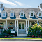 Maison patrimoniale en pierres grises avec des balustrades blanches et des aménagements de fleurs roses et blanches dans les plates-bandes avant et dans des paniers accrochés au toit.